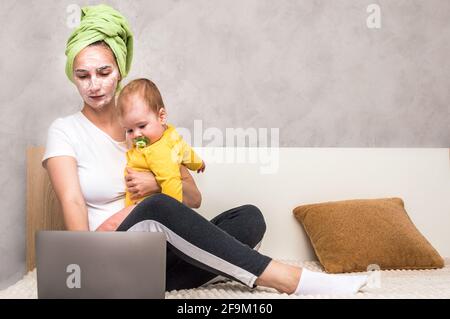 Die junge Frau arbeitet auf einem Bett an einem Laptop und hält ihr Baby in den Armen. Mama ist in Hauskleidung gekleidet, mit einer Maske im Gesicht und einem Handtuch auf den Haaren Stockfoto