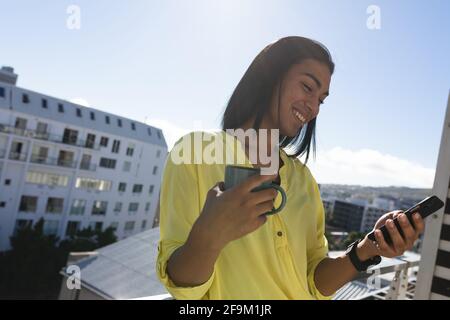 Lächelnde, gemischte Transgender-Frau, die auf der sonnigen Dachterrasse steht Kaffee mit dem Smartphone halten Stockfoto