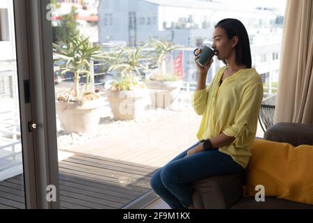 Lächelnde, gemischte Transgender-Frau, die Kaffee trinkt und auf die sonnige Dachterrasse blickt Stockfoto