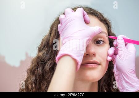 Augenbrauenmeister macht eine Augenbrauenkorrektur für eine junge Frau, die medizinische Handschuhe trägt. Augenbrauen färben. Verfahren, Stufe Stockfoto