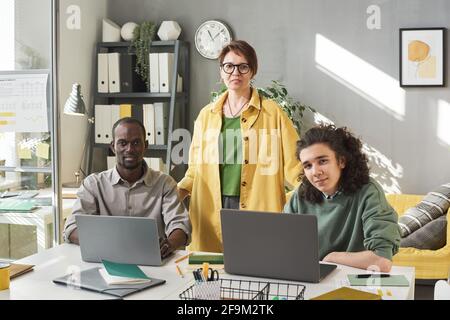 Reife Lehrerin lächelt mit ihren Schülern, die sitzen, vor der Kamera Am Tisch und an Computern im Büro arbeiten Stockfoto