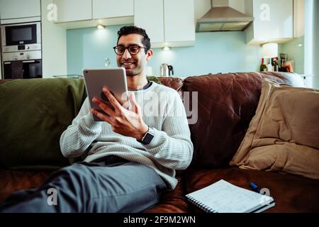 Glücklicher gemischter Geschäftsmann, der von zu Hause aus am Tippen arbeitet Digitales Tablet auf der gemütlichen Couch in der Lounge Stockfoto