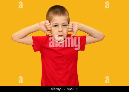 Kind im roten T-Shirt auf gelbem Hintergrund, Junge 5 Jahre alt, traurig. Zeigt die Finger nach unten. Traurige Kinderemotionen Stockfoto