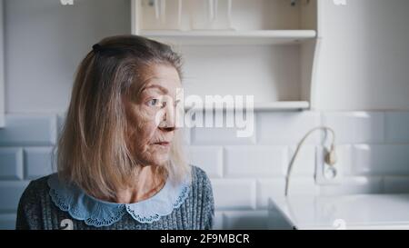 Porträt einer älteren grauhaarigen Dame, die durch das Küchenfenster schaut und denkt. Verletzliche einsame Menschen während des covid Ausbruchs. Hochwertige Fotos Stockfoto