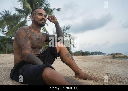 asiatischer Kerl sitzt am Strand mit einem Hund unter Die Palmen Stockfoto