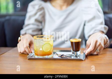 Eine Kombination aus Frischkaffee, Zitrone und Orangensoda auf dem Edelstahlteller auf dem Holztisch vor der Unschärfe-Frau im Café. Stockfoto