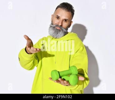 Aktiver alter, brutaler bärtiger Mann mit gelbem Kapuzenpullover, der zwei hält Grüne Hanteln in der Hand auf offener Handfläche und ruft an Beitreten Stockfoto