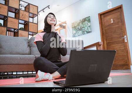 Die Frau in einer Hijab-Sportkleidung sitzt mit gekreuzten Beinen und dehnt sich mit Ihre Hände unter ihrem Kinn und ihr Kopf blickte nach oben Stockfoto