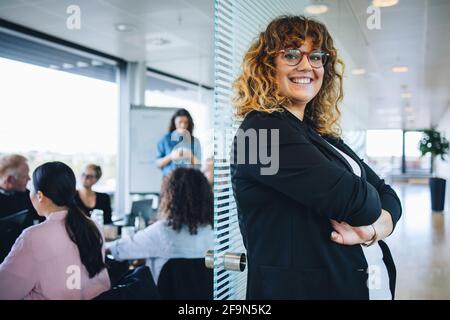 Porträt einer lächelnden Geschäftsfrau, die im Büro mit Kollegen im Besprechungsraum steht. Erfolgreiche weibliche Profikerin mit gekreuzten Armen. Stockfoto
