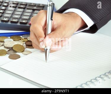 Geschäftsmann Hand hält einen Stift schreiben auf leerem Papier mit Münzen & Rechner beiseite - Business & Financial Konzept Stockfoto