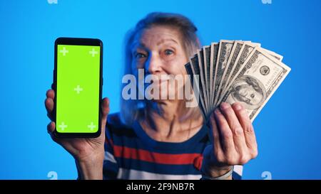Ältere Frau hält Telefon mit grünem Bildschirm in der einen und Geld in der anderen Hand. Lotteriegewinn. Hochwertige Fotos Stockfoto