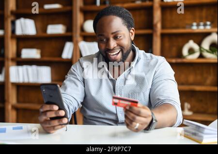 Aufgeregt afroamerikanischen Kerl ist mit einem Smartphone und einer Kreditkarte zum Online-Shopping, glücklich schwarzen Mann bestellt Lebensmittel online, männlich für lang erwartete Kauf auf dem Handy bezahlen. E-Banking Stockfoto