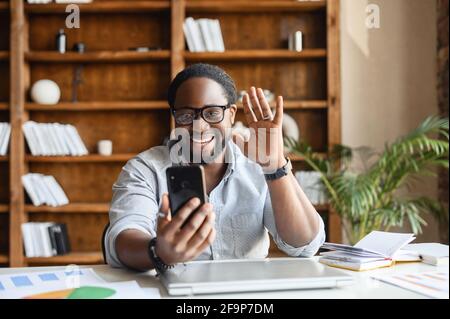 Überglücklich afroamerikanischen Kerl mit einem Smartphone für Videoanruf sitzen im Büro, ein Mixed-Rennen fröhlich jungen Mann winkt hallo, froh, Gesprächspartner, Video-Meeting, virtuelle Konferenz-Konzept zu treffen Stockfoto