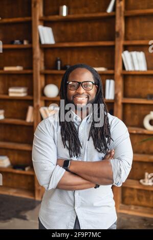 Vertikales Porträt von lächelndem jungen gutaussehenden afroamerikanischen Mann mit Dreadlocks und Brille stehend mit Armen gefaltet und Blick auf die Kamera, optimistischen schwarzen Mann Büroleiter ist bereit zu arbeiten Stockfoto