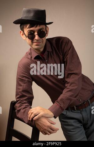 Porträt eines lächelnden Mannes mittleren Alters in braunem Hemd, grauer Hose, Hut und Sonnenbrille, der seinen Ellbogen auf einem schwarzen Stuhl lehnte. Stockfoto