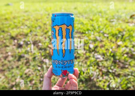 Eine Hand, die die Basis des Monster Energy Drinks vor einem grünen Feld hält. Stockfoto