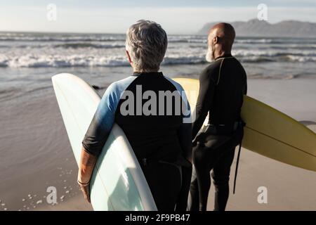 Vielfältiges Seniorenpaar am Strand mit Surfbrettern mit Blick auf das Meer. Gesundheit und Wohlbefinden, aktiver Ruhestand. Stockfoto