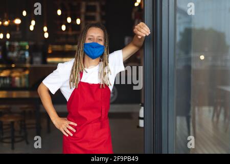 Porträt eines Barista mit gemischter Rasse und Dreadlocks mit Gesichtsmaske, die sich in der Tür des Cafés lehnt. Unabhängiges kleines Unternehmen während des Coronavirus-Covids Stockfoto