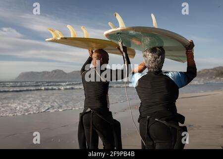 Ältere afroamerikanische Paar mit Surfbrett auf dem Kopf am Strand. Reise Urlaub Ruhestand Lifestyle-Konzept Stockfoto