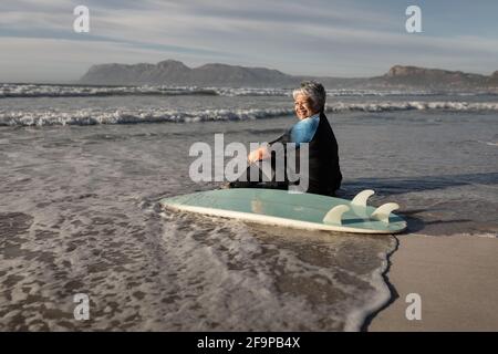 Ältere afroamerikanische Frau mit Surfbrett lächelnd, während sie am Strand sitzt. Reise Urlaub Ruhestand Lifestyle-Konzept Stockfoto