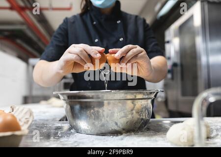 Mittelteil des professionellen Küchenchefs, der Eier mit Gesichtsmaske bricht. Während der Coronavirus-Pandemie 19 in einer geschäftigen Restaurantküche arbeitet. Stockfoto