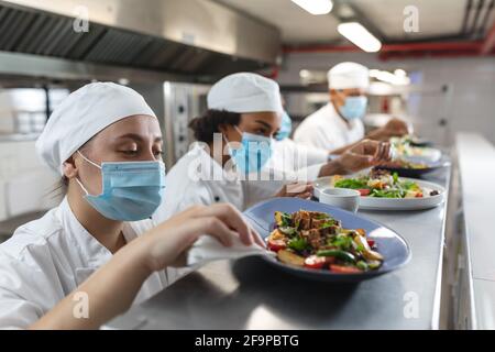 Diverce Rennen männlichen und weiblichen professionellen Köche verschenken Gerichte mit Gesichtsmasken. Arbeiten in einer geschäftigen Restaurantküche während Coronavirus covid 1 Stockfoto