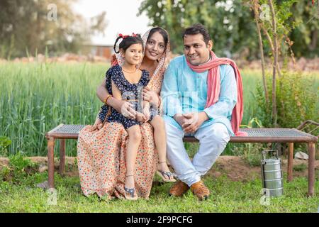 Glückliche indische Bauernfamilie, die auf einem traditionellen Bett auf einem landwirtschaftlichen Feld im Dorf sitzt, Junges Paar mit ihrer Tochter in traditionellem Outfit. Stockfoto