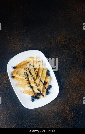 Hausgemachte Crepes, leckere dünne Pfannkuchen mit Schokolade und Beeren auf einem grauen Teller zum Frühstück auf dunklem Hintergrund. Traditionelle russische Küche. Stockfoto