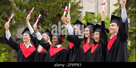 Glückliche multirassische Studenten in Abschlusskostümen, die Diplome hochziehen, Panorama Stockfoto