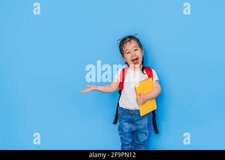 Überrascht schulmädchen hält gelbe Buch in einer Hand und zeigt auf die Seite vor blauem Hintergrund mit der anderen. Zurück zur Schule im Jahr 2021. Homescho Stockfoto