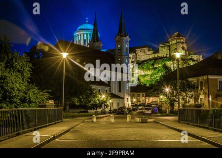 Nachtansicht einer schmalen Straße in der ungarischen Stadt Esztergom mit der Kirche des heiligen Ignac und der Basilika auf dem Hügel. Stockfoto