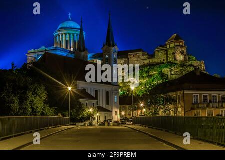 Nachtansicht einer schmalen Straße in der ungarischen Stadt Esztergom mit der Kirche des heiligen Ignac und der Basilika auf dem Hügel. Stockfoto