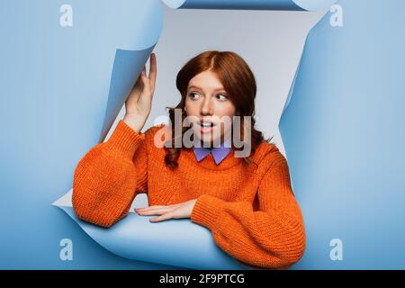 Überrascht junge Frau in orange Pullover Blick weg auf blau Gerippte Hintergrundfarbe Stockfoto