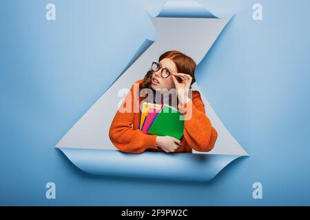 Rothaarige Schülerin in orangefarbenem Pullover und Brille mit Notizbüchern Blauer zerrissener Papierhintergrund Stockfoto