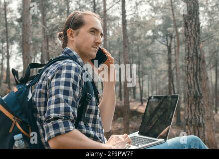 Junger Mann, der am Wald sitzt und auf dem Smartphone spricht und arbeitet Auf einem Laptop Stockfoto