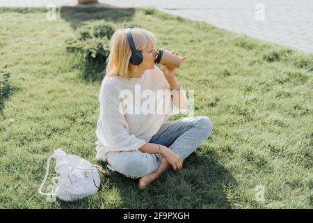 Junge Frau mit Kopfhörern, die draußen auf dem Gras sitzt parken Stockfoto