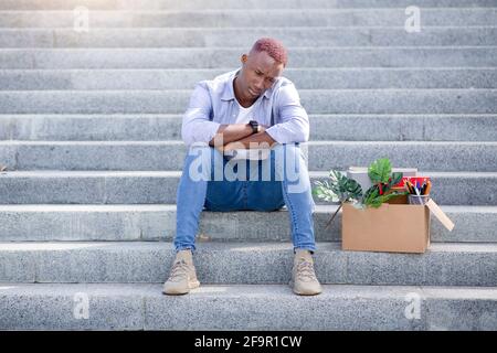 Konkurs und Arbeitslosigkeit. Gestresster schwarzer Kerl mit persönlichem Zeug, der sich nach dem Verlust der Arbeit auf der Treppe draußen verärgert fühlt Stockfoto