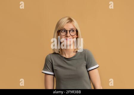 Nachdenkliche Frau in einer Brille, die über Fragen nachdenkt, das Kinn berührt und wegschaut, beiger Hintergrund Stockfoto