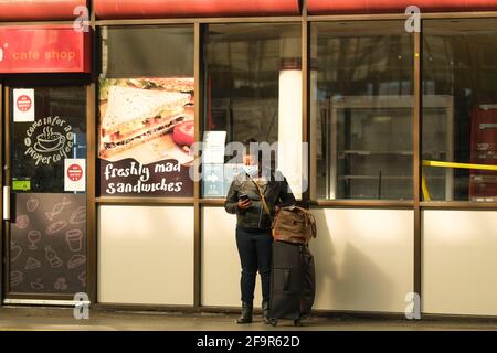 Frau mit Koffern, die eine Gesichtsmaske tragen und ihr Telefon überprüfen, während sie am Bahnhof Preston auf einen Zug wartet. Stockfoto