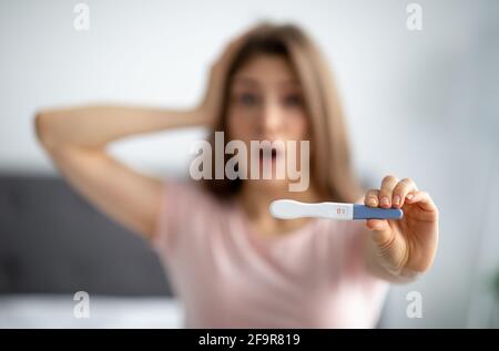 Schockierte junge Frau, die vor der Kamera einen positiven Schwangerschaftstest zeigte und sich vor Angst den Kopf packte Stockfoto