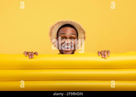 Millennial Black Guy in Strohhut und Sonnenbrille versteckt sich dahinter Aufblasbare Lilo auf gelbem Studiohintergrund Stockfoto