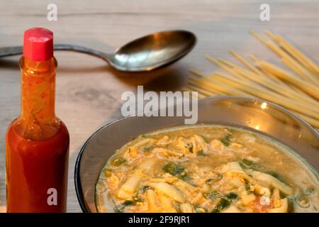 Würzige asiatische Nudelsuppe in einer Glasschüssel, Tabasco-Sauce und ungekochte Nudel auf einem Holzbrett serviert Stockfoto