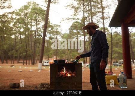 Junger Mann, der das Abendessen am Lagerfeuer neben seinem vorbereitet Campingplatz Stockfoto