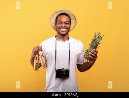 Fröhlicher schwarzer Kerl im Sommer-Outfit mit Kamera, die Bier hält Flaschen und Ananas auf gelbem Studiohintergrund Stockfoto