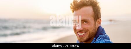 Lächelnd hübscher junger Mann Schönheitsporträt am Strand bei Sonnenuntergang Blick auf die Kamera lachend, gesundes Grinsen -Panorama-Banner Gesicht von glücklichem Model in Handtuch. Stockfoto