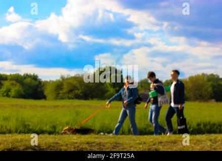 Verschwommener Hintergrund einer Gruppe von Menschen mit einem Corgi-Hund, der an einem sonnigen Tag in einem Frühlingspark läuft. Verschwommene Aufnahme. Grünes Gras und blauer Wolkenhimmel. Stockfoto