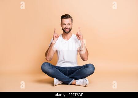 Porträt eines netten, fröhlichen Typen, der in Lotos auf dem Boden sitzt Pose zeigt den Zeigefinger nach oben, die Anzeige ist auf beige isoliert Farbiger Hintergrund Stockfoto