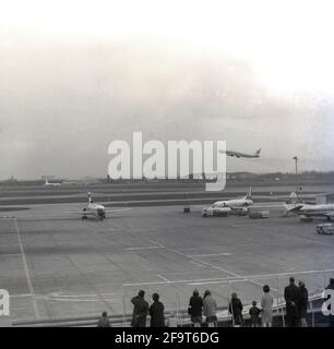 1967, historisch, am flughafen heathrow geparkte Düsenflugzeuge und ein Flugzeug, das in das Passagierausschiffungsgebiet, London, England, Großbritannien, einsteigt. In der Ferne fliegt ein Düsenflugzeug ab. Ein Jahr zuvor wurde der Flughafen London 1966 nach dem ursprünglichen ländlichen Dorf Heath Row, das das Land für den Flughafen lieferte, in Heathrow umbenannt. Das Dach des Europa Terminals war eine beliebte Aussichtsplattform. In der Ferne ein Schild mit der Aufschrift „Hunting“, das ein Zeichen für den alten Hangar des Jagdclans war, der von Field Aircraft Services Ltd verwendet wurde, obwohl das alte Zeichen erhalten blieb. Stockfoto