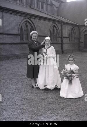 1956, draußen auf dem Gelände einer Kirche, eine stolze Mutter mit ihrer süßen jungen Tochter, die ihre May Queen-Krone und einen Blumenstrauß trägt, mit einem kleinen Mädchen neben ihr, das ebenfalls einen Blumenstrauß hält, England, Großbritannien. Der Maifeiertag ist ein uraltes Fest, das die Ankunft des Frühlings feiert, einschließlich der Krönung einer Maikönigin und des Tanzens um einen Maypole, Aktivitäten, die seit Jahrhunderten in England stattfinden. Die May Queen, die ihre Krone trug, wurde von den Mädchen der Region ausgewählt, die die Parade zum 1. Mai anführen sollten, und im Norden Englands führten die Church Sunday Schools die Organisation oft an Stockfoto
