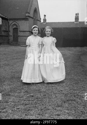 1956, historisch, draußen auf dem Gelände einer Kirche, stehend für ein Foto, zwei junge Mädchen in ihren hübschen Kleidern, die sie bei der traditionellen May Queen Prozession, England, Großbritannien, tragen werden. Der Maifeiertag ist ein uraltes Frühlingsfest, zu dem Morris Dancing, die Krönung einer Maikönigin, und das Tanzen um einen Maypole gehören, der seit Jahrhunderten in England stattfindet. Die May Queen, die eine Krone trägt, wurde von den Mädchen der Region ausgewählt, die die Parade zum 1. Mai anführen sollten, und im Norden Englands führten die Church Sunday Schools die Organisation des Tages an. Stockfoto